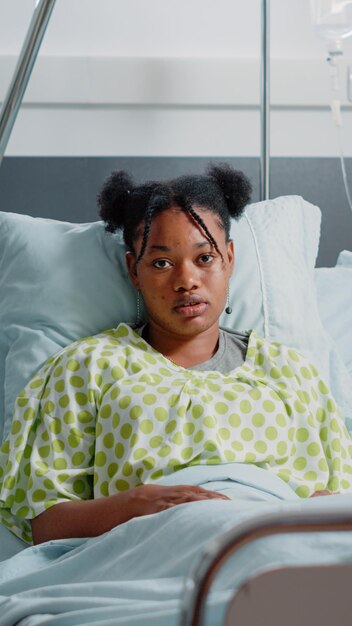 Portrait of young patient in bed with IV drip bag, waiting on doctor to give medical assistance and cure disease. Sick woman sitting in hospital ward with heart rate monitor to heal