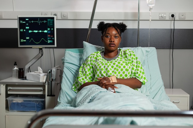 Portrait of young patient lying in bed during clinical examination recovering after medical surgery in hospital ward. Sick woman looking into camera waiting for healthcare treatment