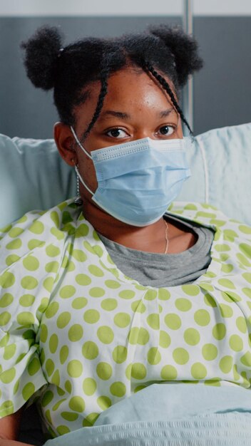 病棟でフェイスマスクを着用して病気の若い患者の肖像画。コロナウイルスのパンデミック中に医師からの病気に対する医療援助を待っているベッドで病気の女性。