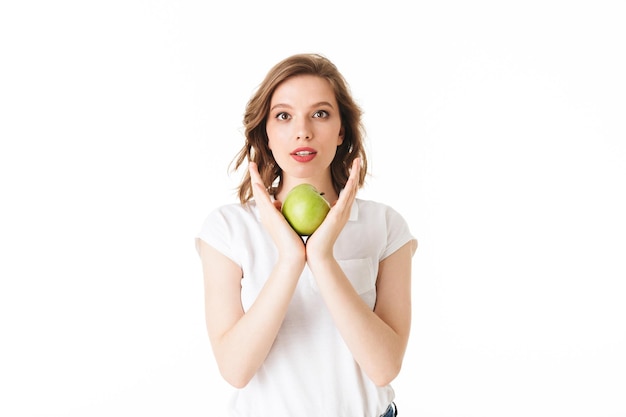 Portrait of young pensive lady standing with green apple in hand and dreamily looking in camera on white background isolated