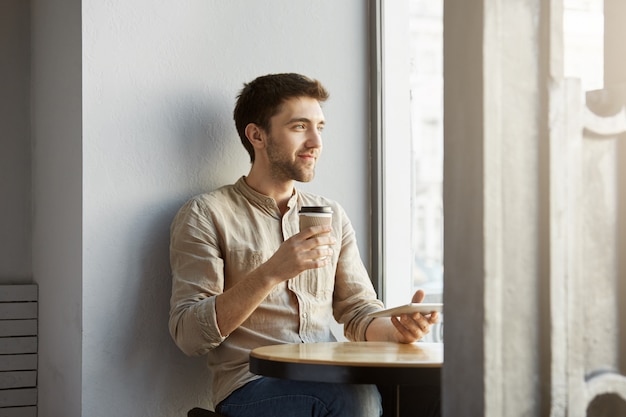 Free photo portrait of young perspective male freelance designer sitting in cafeteria, looking aside being satisfied with his new project.