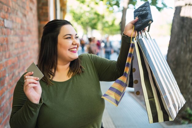 Portrait of young plus size woman holding a credit card and shopping bags outdoors on the street. Shopping and sale concept.