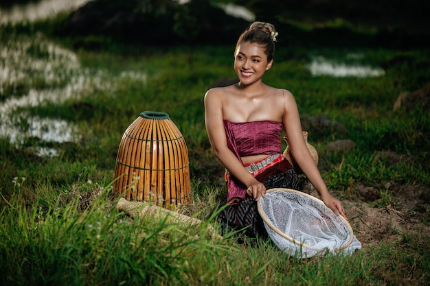 Foto gratuita ritratto giovane bella donna asiatica in bellissimi abiti tradizionali tailandesi al campo di riso, seduta vicino all'attrezzatura da pesca