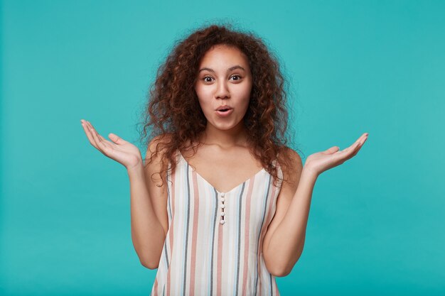 Portrait of young pretty brown haired curly female raising perplexedly hands while looking positively, isolated on blue in summer top
