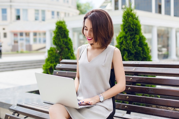 A portrait of a young pretty brunette girl  sitting on the bench in city. She wears gray and black dress. She is typing on laptop.