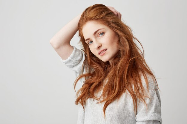 Portrait of young pretty ginger girl with freckles smiling touching hair .