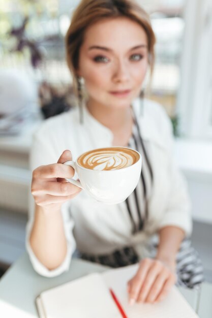 Portrait of young pretty girl with blond hair sitting in restaurant with notebook on table and drinking coffee. Close up woman hand holding cup of cappuccino in coffee shop