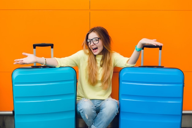 Portrait of young pretty girl with long hair in black glasses sitting on orange background between two suitcases. she has long hair and yellow sweater with jeans. she is smiling to the camera