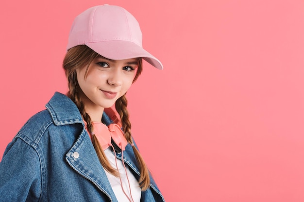 Free photo portrait of young pretty joyful girl with two braids in denim jacket and cap with headphones on neck happily looking in camera over pink background isolated