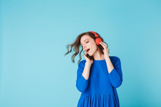 Portrait of young pretty lady in blue dress dancing while listening music in headphones on over pink background