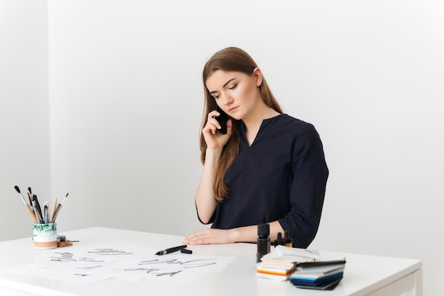 Portrait of young pretty lady sitting at the white desk and talking on her cellphone while thoughtfully looking aside isolated