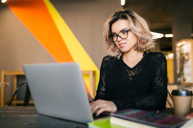 Free photo portrait of young pretty smiling woman sitting at table in black shirt working on laptop in co-working office