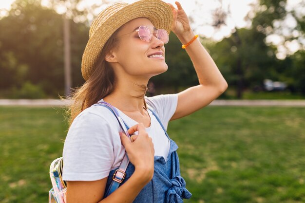 Portrait of young pretty smiling woman in straw hat and pink sunglasses walking in park, summer fashion style, colorful hipster outfit