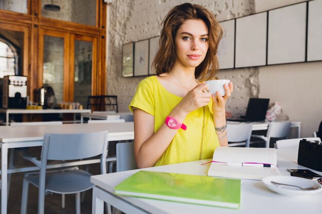Portrait of young pretty woman sitting at table in cafe drinking coffee, holding cup in hands, student learning, education