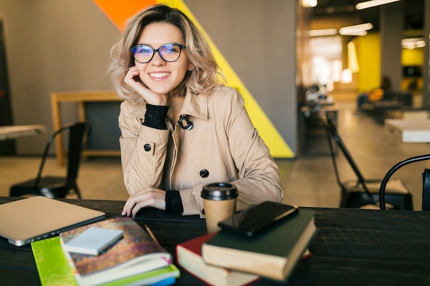 Foto gratuita ritratto di giovane donna graziosa che si siede al tavolo in trench lavorando sul portatile in ufficio di co-working, con gli occhiali, sorridente, felice, positivo, sul posto di lavoro