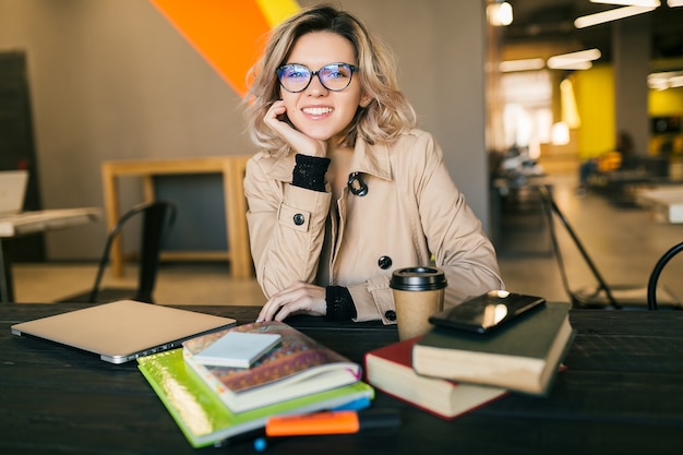 Ritratto di giovane donna graziosa che si siede al tavolo in trench, lavorando sul computer portatile in ufficio di co-working