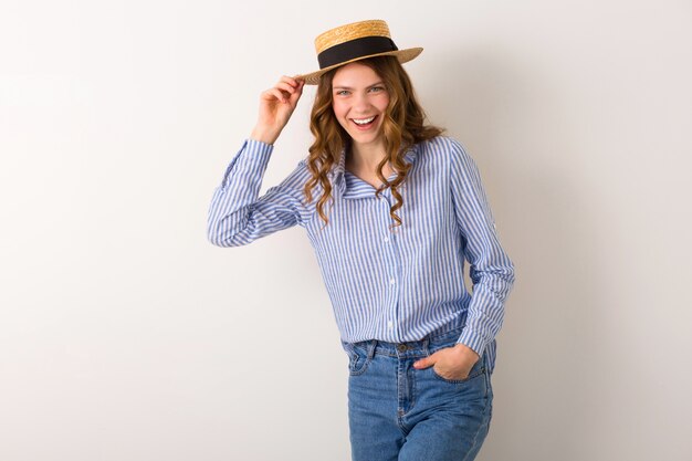 Портрет молодой красивой женщины с соломенной шляпой, джинсами, синей хлопковой рубашкой, позирует на белой стене