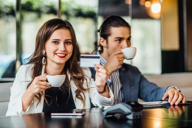 Portrait of young prety woman holding credit card at hands while man drinking coffee.