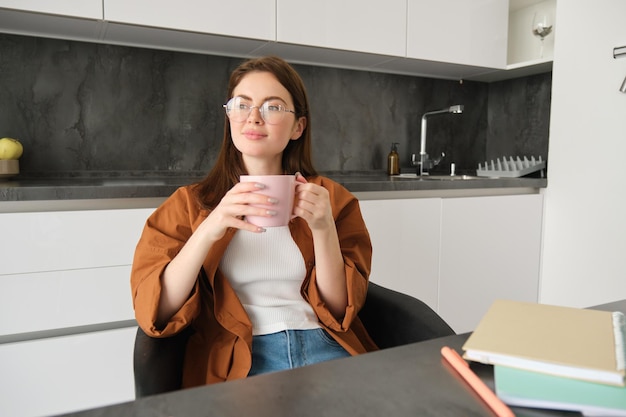 Free photo portrait of young professional girl freelancer sitting in kitchen and enjoying cup of coffee sitting