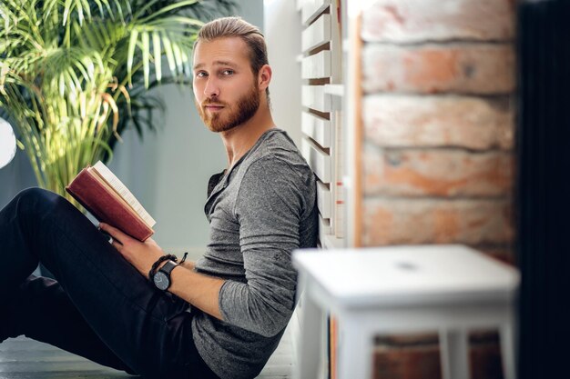 Portrait of young redhead bearded male holding a book in a room with green plants.