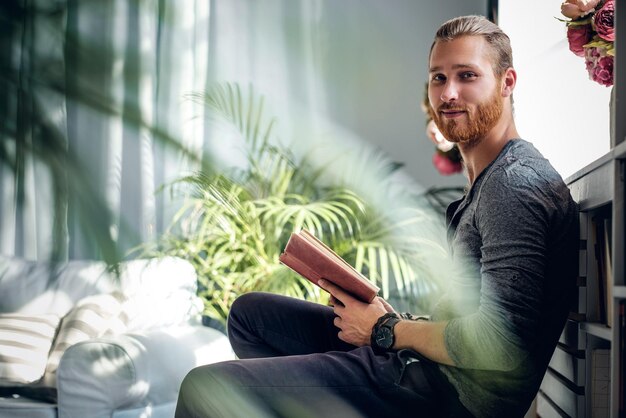 Portrait of young redhead bearded male holding a book in a room with green plants.
