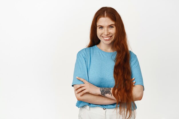 Portrait of young redhead confident female student, ginger girl with long natural hair, holding arms crossed on chest, smiling satisfied, standing on white.