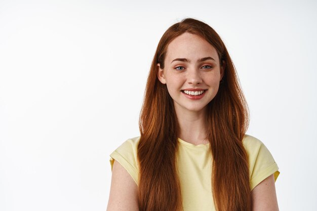 Portrait of young redhead girl face, long natural foxy hair and white smile, looking happy and carefree, standing relaxed in t-shirt against white background.