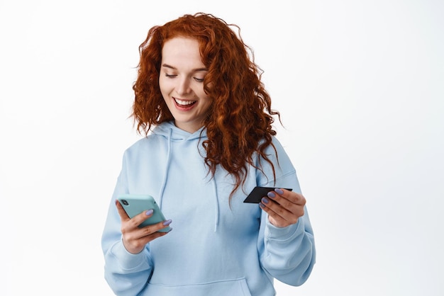 Ritratto di giovane ragazza rossa che effettua l'acquisto paga con smartphone e carta di credito in plastica guardando il telefono cellulare inserire le cifre della carta sullo sfondo bianco