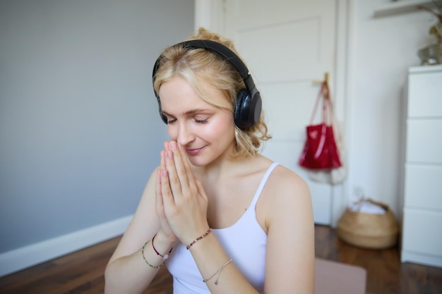 Free photo portrait of young relaxed woman sits in room in headphones clasp hands together meditated listens to