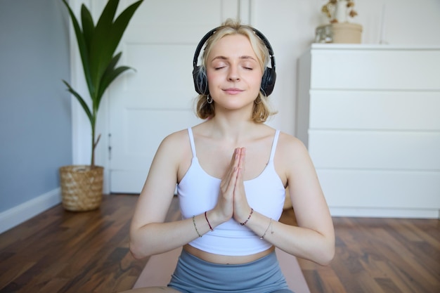 Free photo portrait of young relaxed woman sits in room in headphones clasp hands together meditated listens to