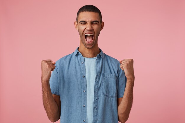 Portrait of young screaming young attractive dark skinned guy in checkered shirt, with fists up and closed eyes, rejoices his favorite teams, stands over pink background
