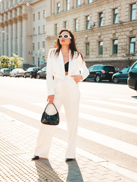 Portrait of young sexy modern businesswoman model. hot beautiful woman in white suit posing on the street background. fashion female walking outdoors in sunglasses