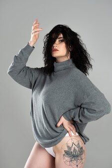 Portrait of young and sexy woman with black curly hair wearing turtleneck jumper posing on gray background in studio