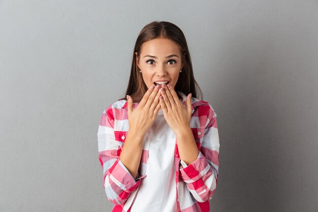 Portrait of young shocked brunette girl covering her mouth in surprise