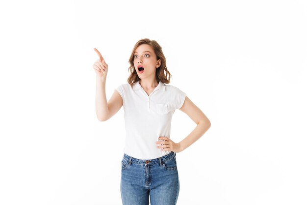 Портрет молодой шокированной дамы, стоящей в футболке и джинсах и изумленно смотрящей в сторону на белом фоне изолированы