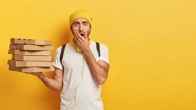 Portrait of young shocked male delivery worker holds stack of pizza boxes, dressed casually, covers opened mouth, stands against yellow wall