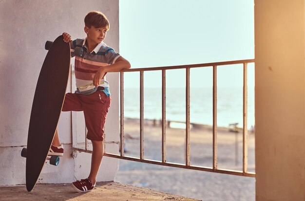 Портрет молодого фигуриста, одетого в футболку и шорты, опирающегося на ограждение и держащего скейтборд на фоне морского побережья на закате.