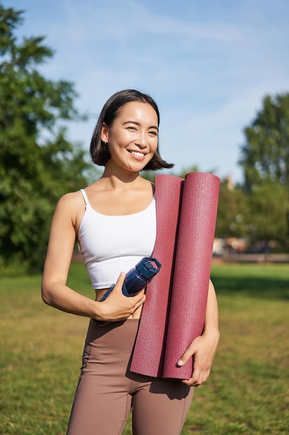 Free photo portrait of young slim and healthy korean girl doing workout in park standing with water bottle and