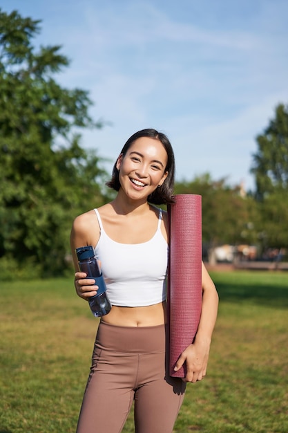 水のボトルと立っている公園でトレーニングを行う若いスリムで健康な韓国の女の子の肖像画と