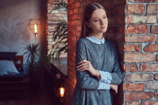 Portrait of a young slim sensual girl in a gray dress leaning against a brick wall with a crossed arms.