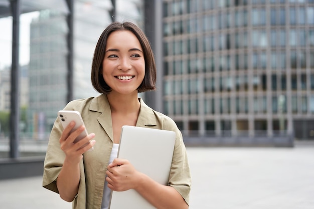 ラップトップとスマートフォンで仕事に行く通りを歩く若い笑顔のアジアの女性の肖像画