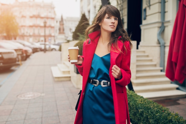 Портрет молодой улыбающейся красивой стильной женщины, идущей по улице города в красном халате, пить кофе
