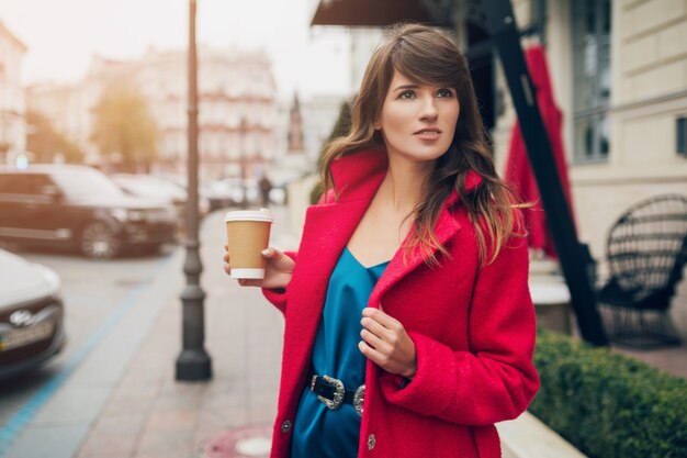 コーヒーを飲む赤いコートで街を歩いている若い笑顔の美しいスタイリッシュな女性の肖像画