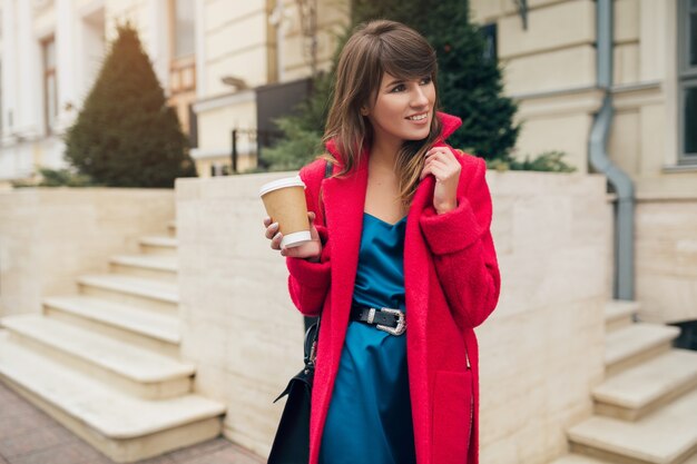 Portrait of young smiling beautiful stylish woman walking in city street in red coat drinking coffee