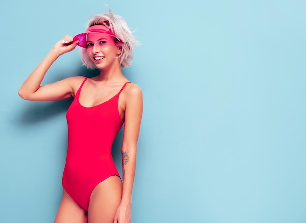Portrait of young smiling blond model in summer swimwear red bathing suit and transparent visor cap Sexy carefree woman having fun and going crazy Female posing near blue wall in studio