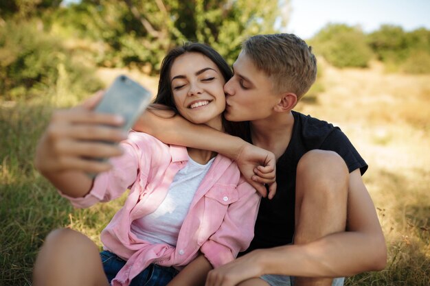 Portrait of young smiling couple sitting on lawn in park and making selfie. Beautiful couple taking photos on cellphone frontal camera