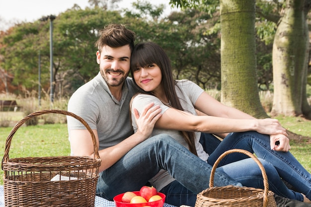 Portrait of young smiling couple sitting in the park with fruits and picnic basket