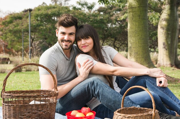 Портрет молодой улыбается пара, сидя в парке с фруктами и корзины для пикника