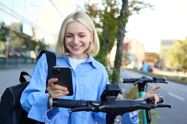 Foto gratuita ritratto di una giovane modella sorridente che affitta uno scooter elettrico usando un telefono cellulare per scansionare qr