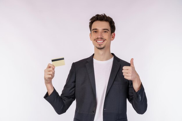 Портрет молодого улыбающегося красивого бизнесмена в повседневной одежде, показывающего кредитную карту и большой палец вверх на белом фоне
